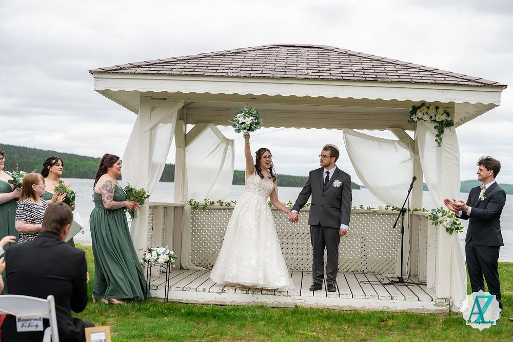 Wedding couple celebrates with guests under a decorated white gazebo surrounded by flowers on a cloudy day.