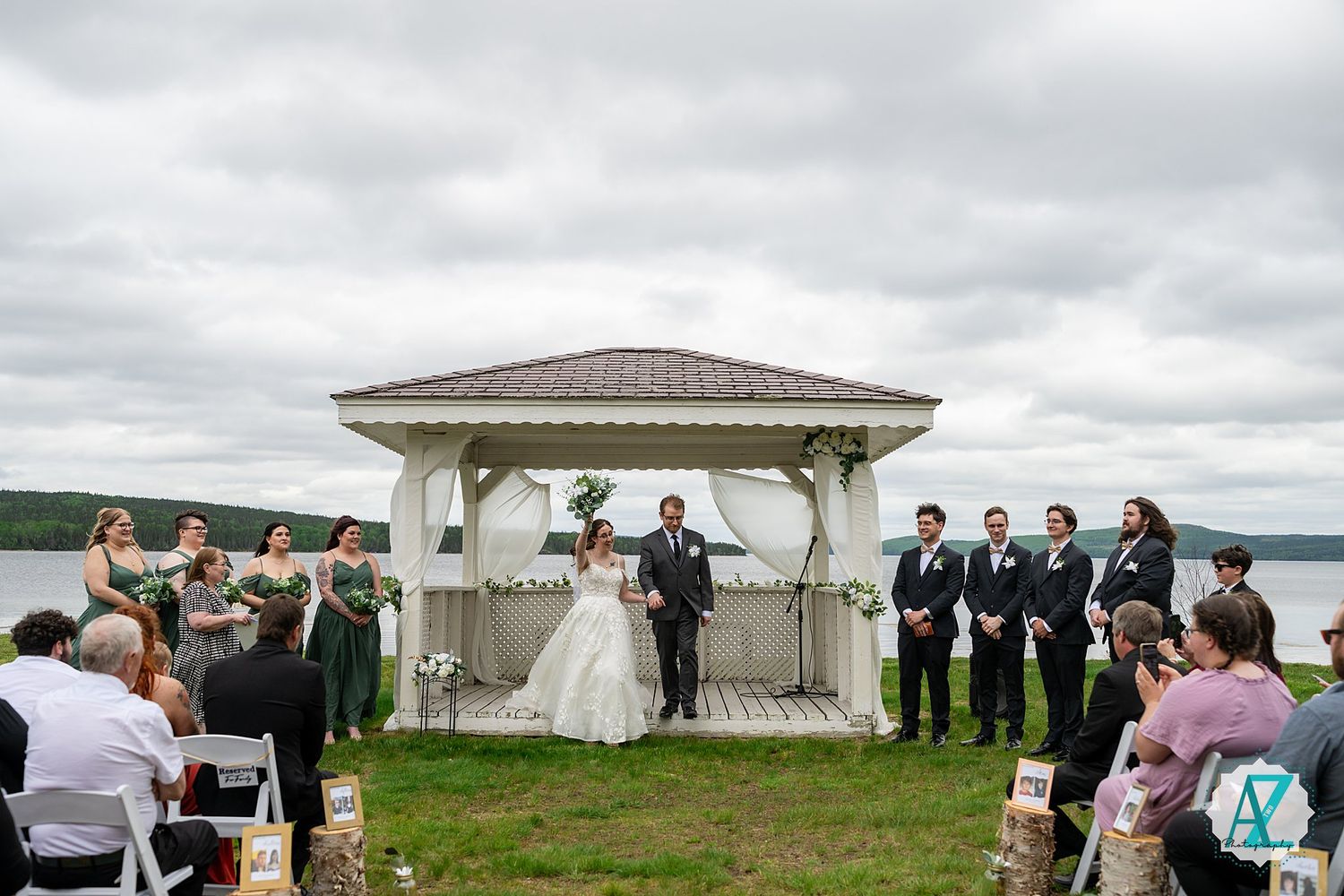 Outdoor wedding ceremony taking place under a white gazebo overlooking a serene waterfront on a cloudy day.