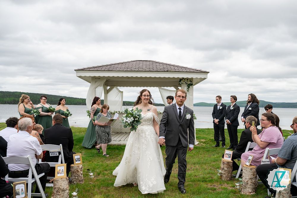 Small outdoor wedding ceremony celebration near a waterfront gazebo on a cloudy day with guests seated on white chairs.