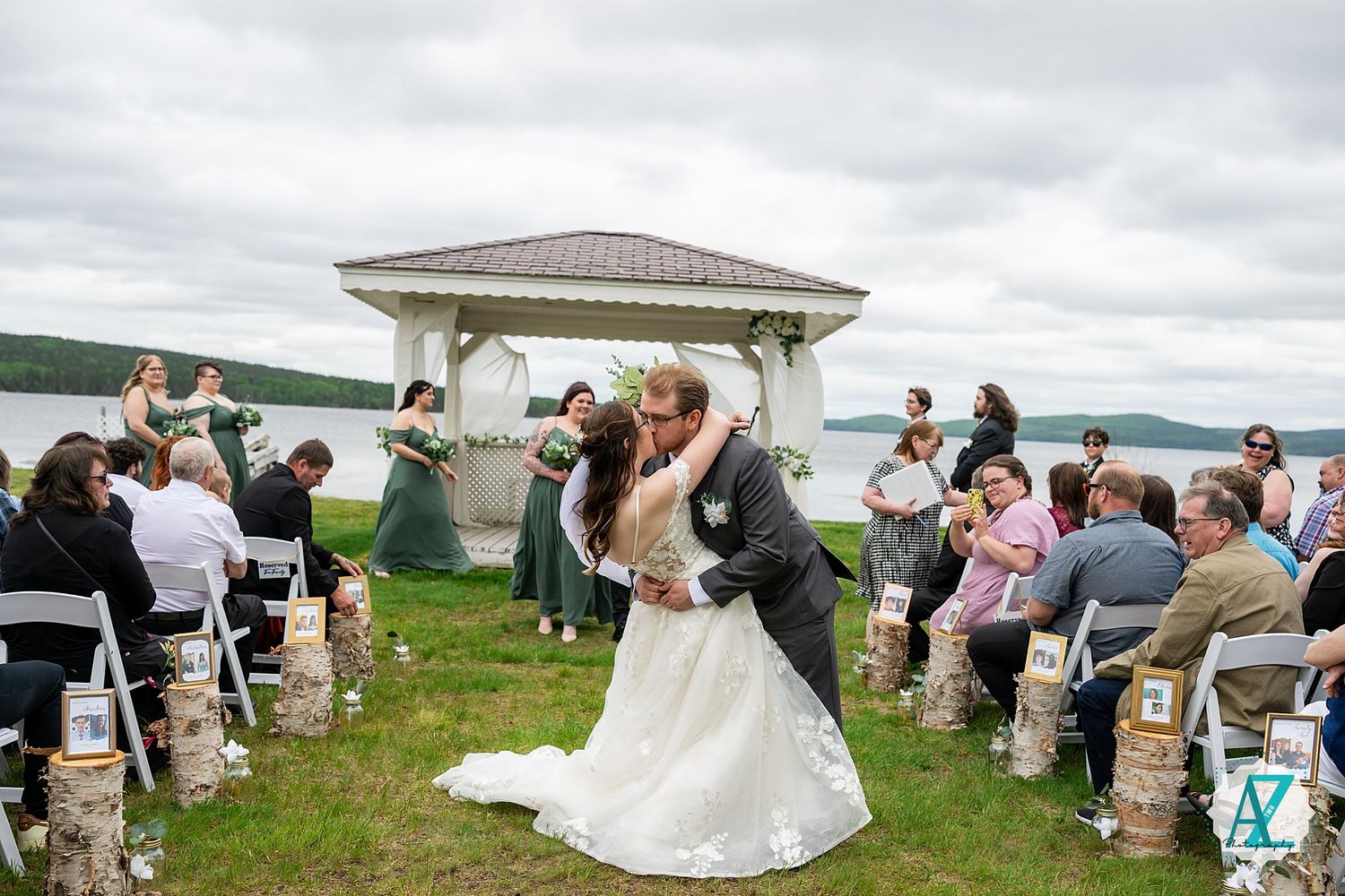 A couple shares their first kiss during a lakeside wedding ceremony under a white gazebo with guests looking on.