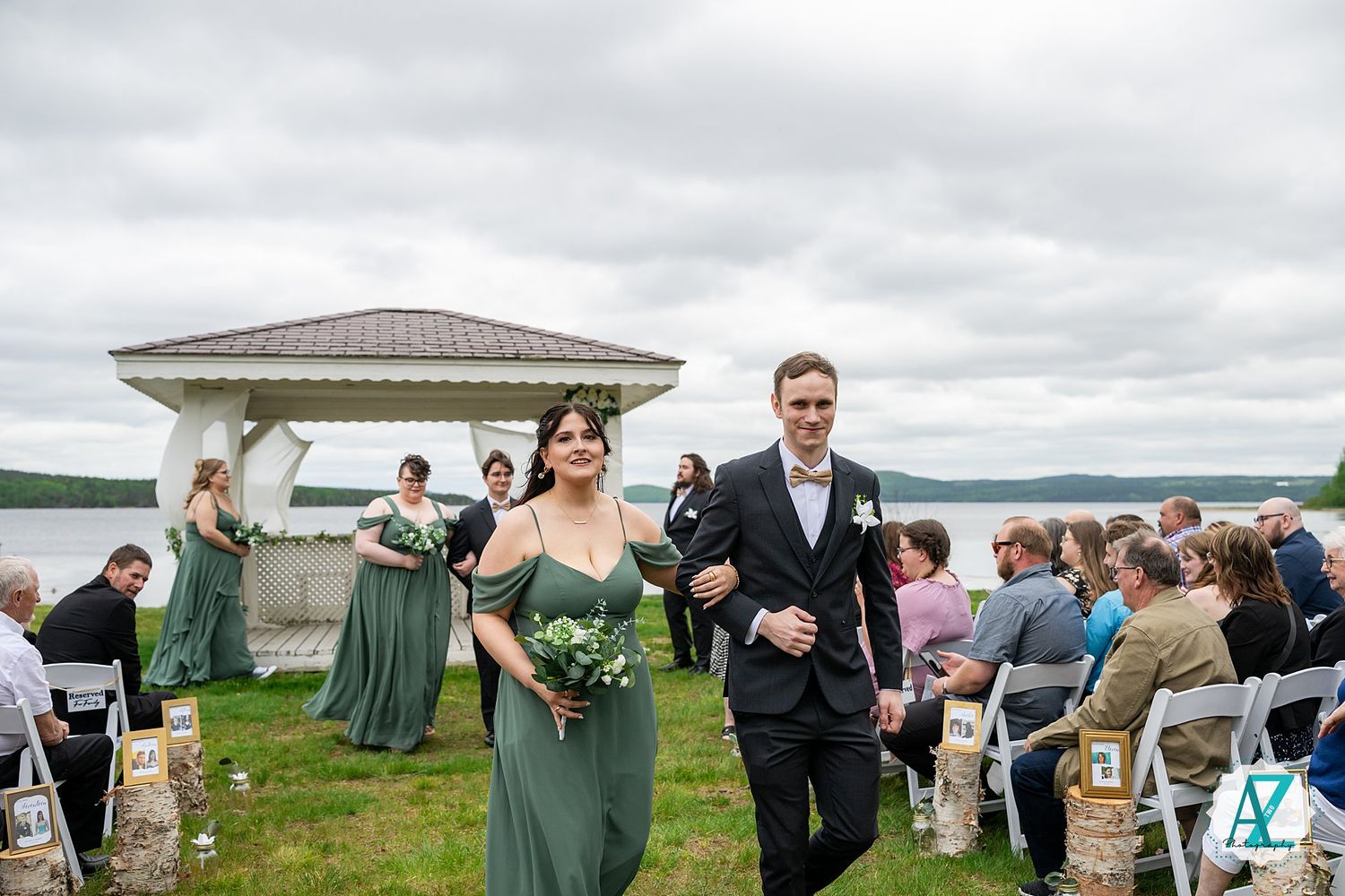 Wedding ceremony participants walk down the aisle at an outdoor gazebo venue overlooking a scenic waterfront.