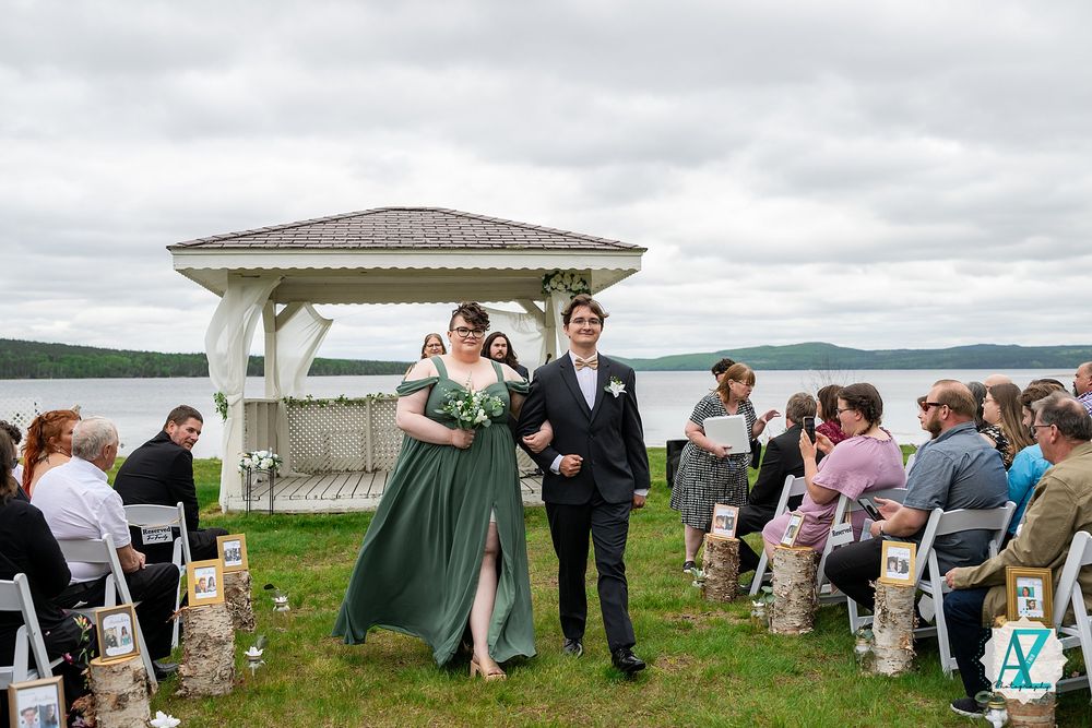 A lively outdoor wedding ceremony takes place under a white gazebo overlooking a scenic lake on a cloudy day.