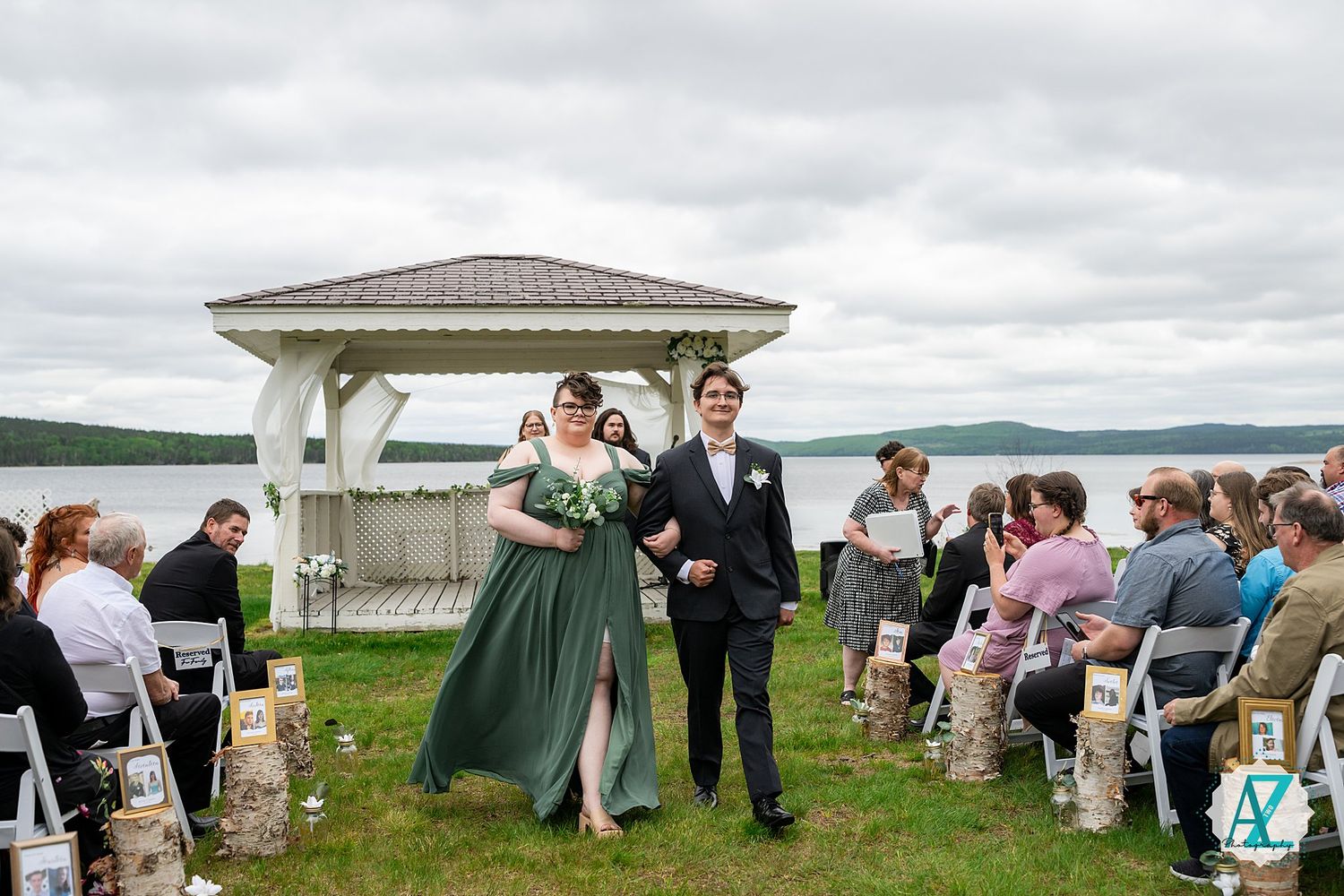 A lively outdoor wedding ceremony takes place under a white gazebo overlooking a scenic lake on a cloudy day.