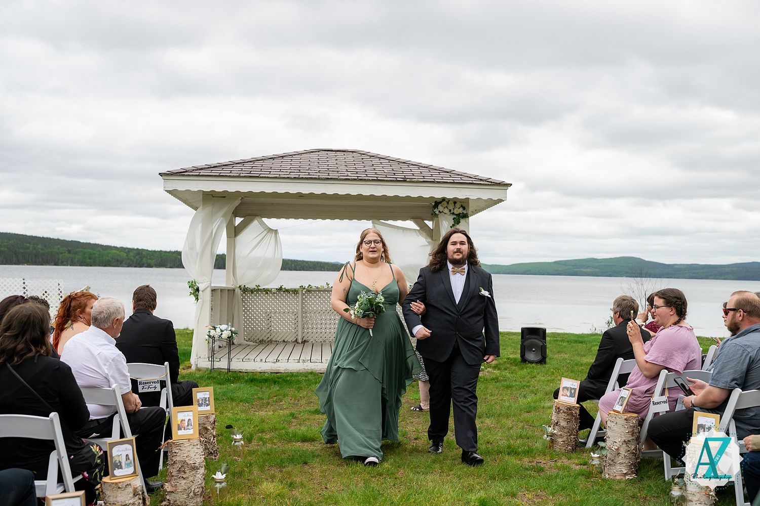 Wedding ceremony on grassy lawn with white gazebo overlooking a serene lake under cloudy skies.