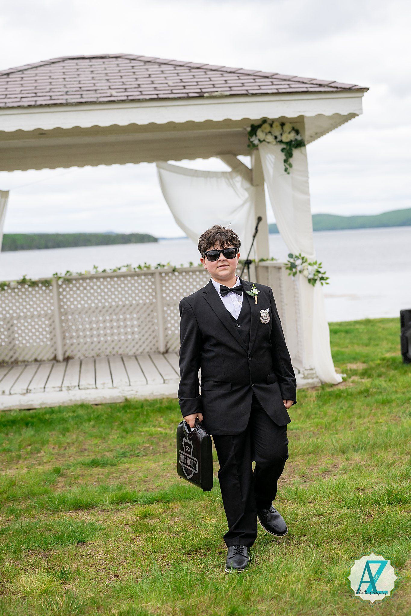A young attendant in a black suit and sunglasses carries a sign while walking beside a decorated gazebo by the water.
