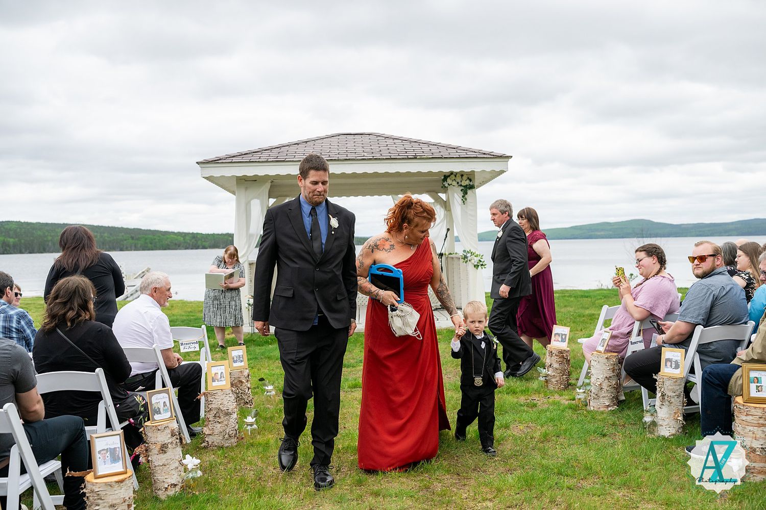 Wedding guests celebrate at an outdoor lakeside ceremony with green lawn and cloudy skies in the background.