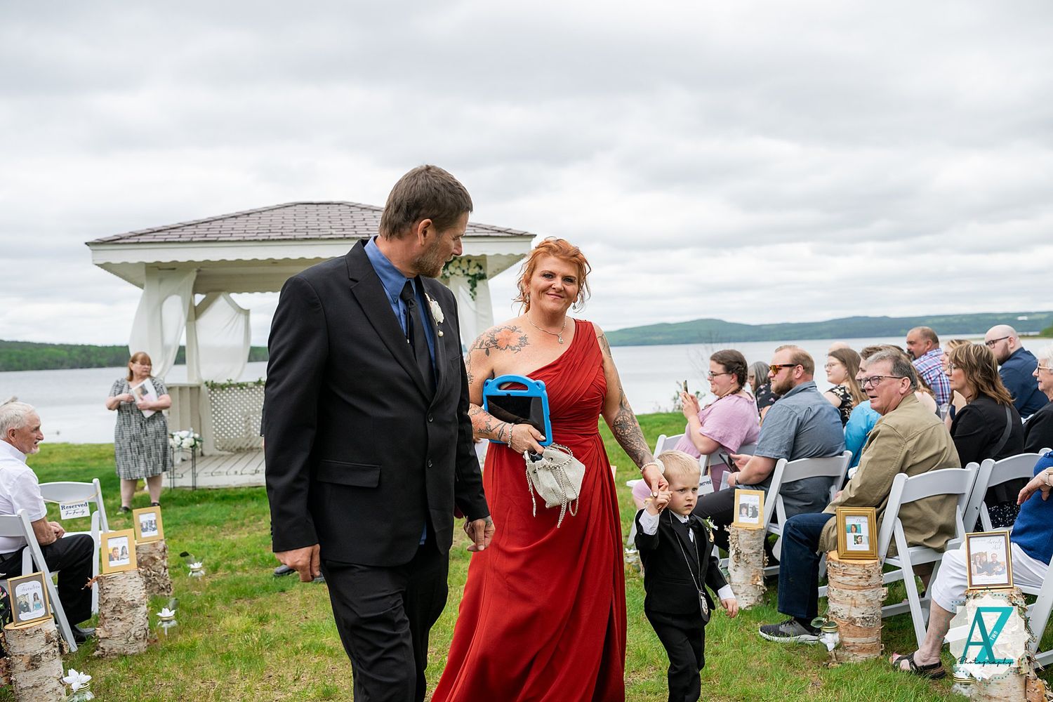 Two wedding guests in formal attire walk past a white gazebo during an outdoor ceremony by a lake under cloudy skies.