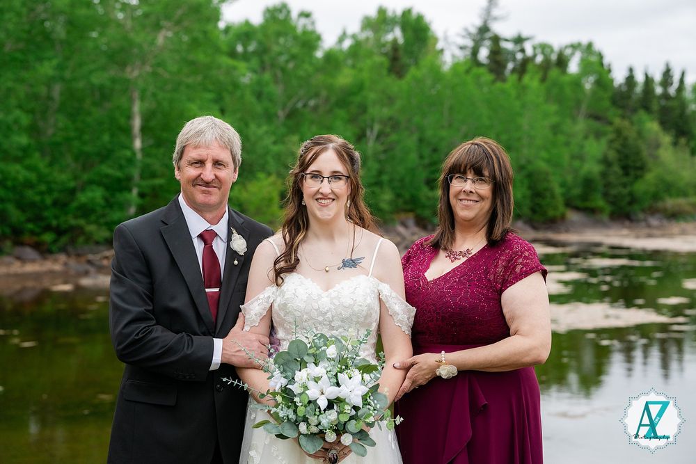 A bride with a white wedding dress and floral bouquet poses near a lake with two others dressed in formal attire.