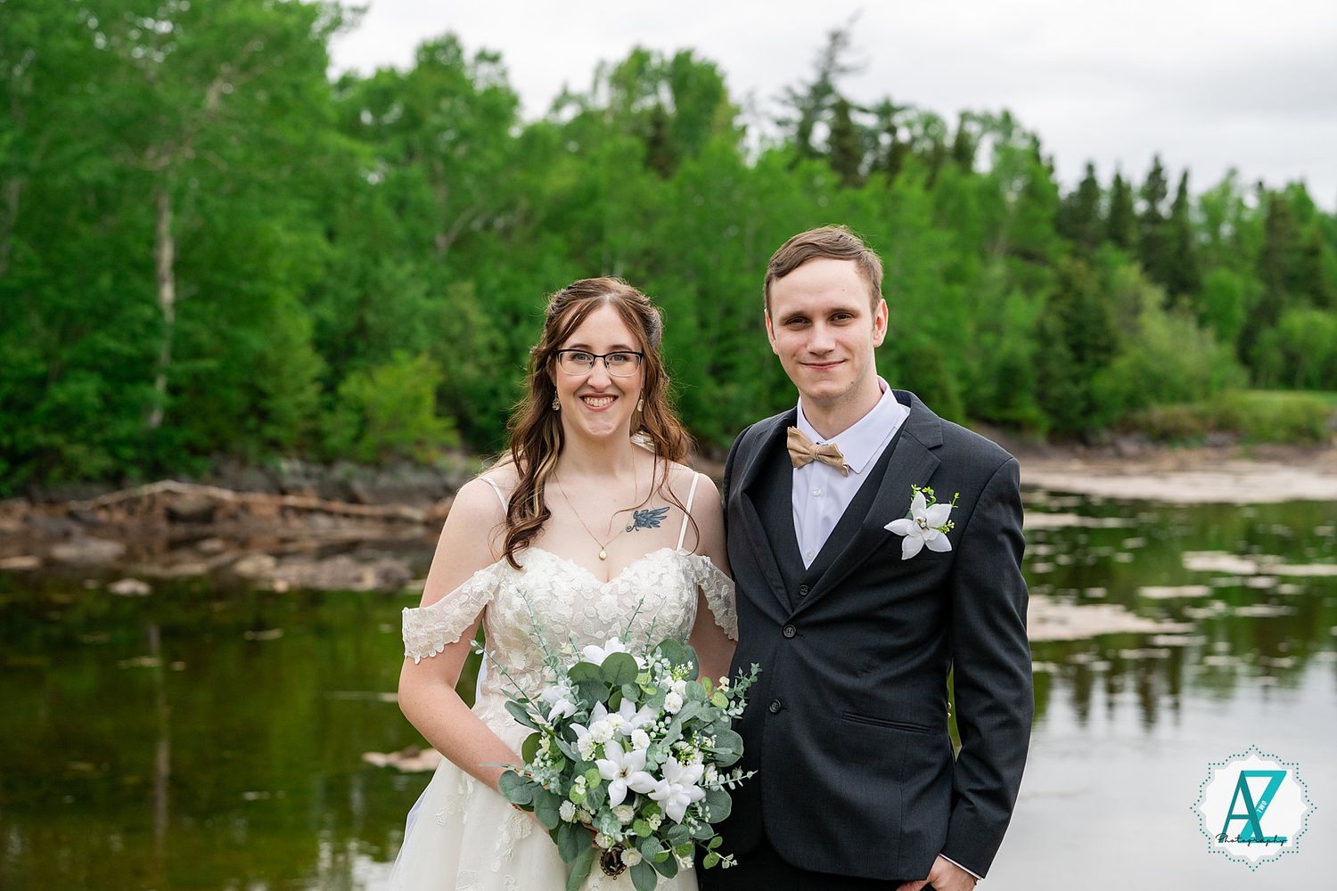 Wedding couple stands near a lakeside with green trees, holding a white floral bouquet against a scenic natural backdrop.