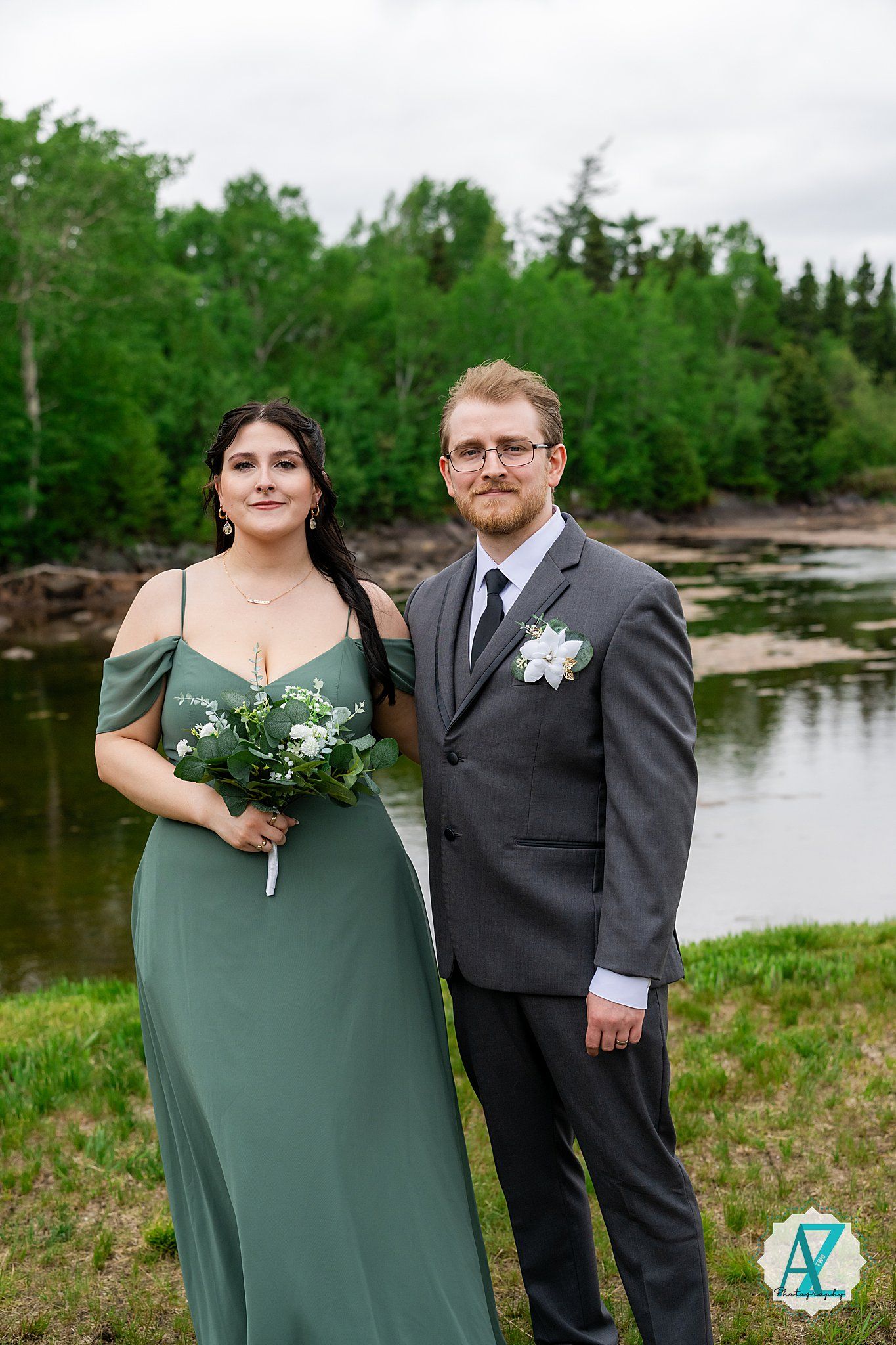 A bridesmaid in a sage green dress and groomsman in grey suit pose by a scenic riverside with trees in background.