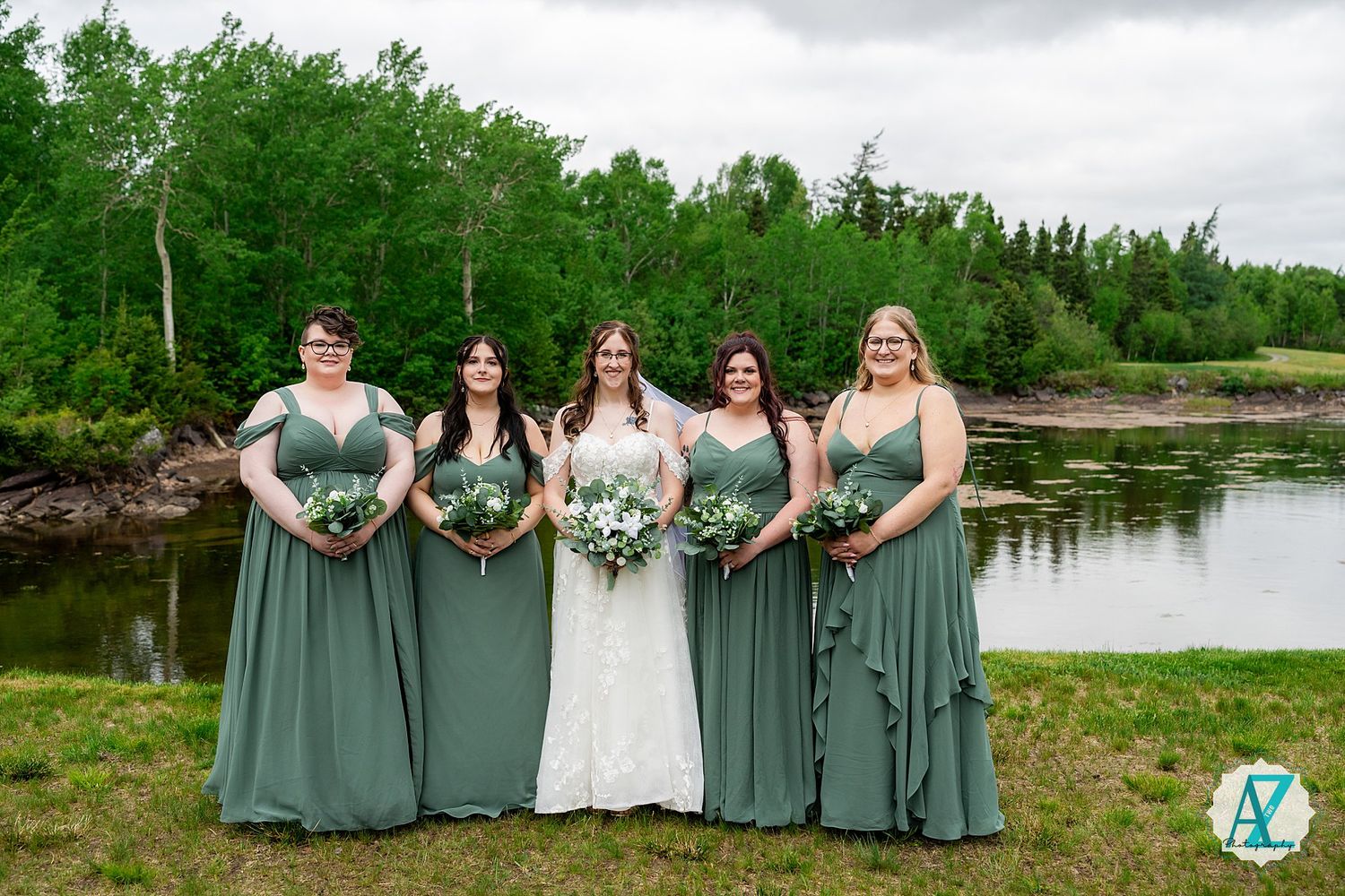 Wedding party members in flowing sage green dresses stand beside bride in white gown near tranquil waterfront setting.