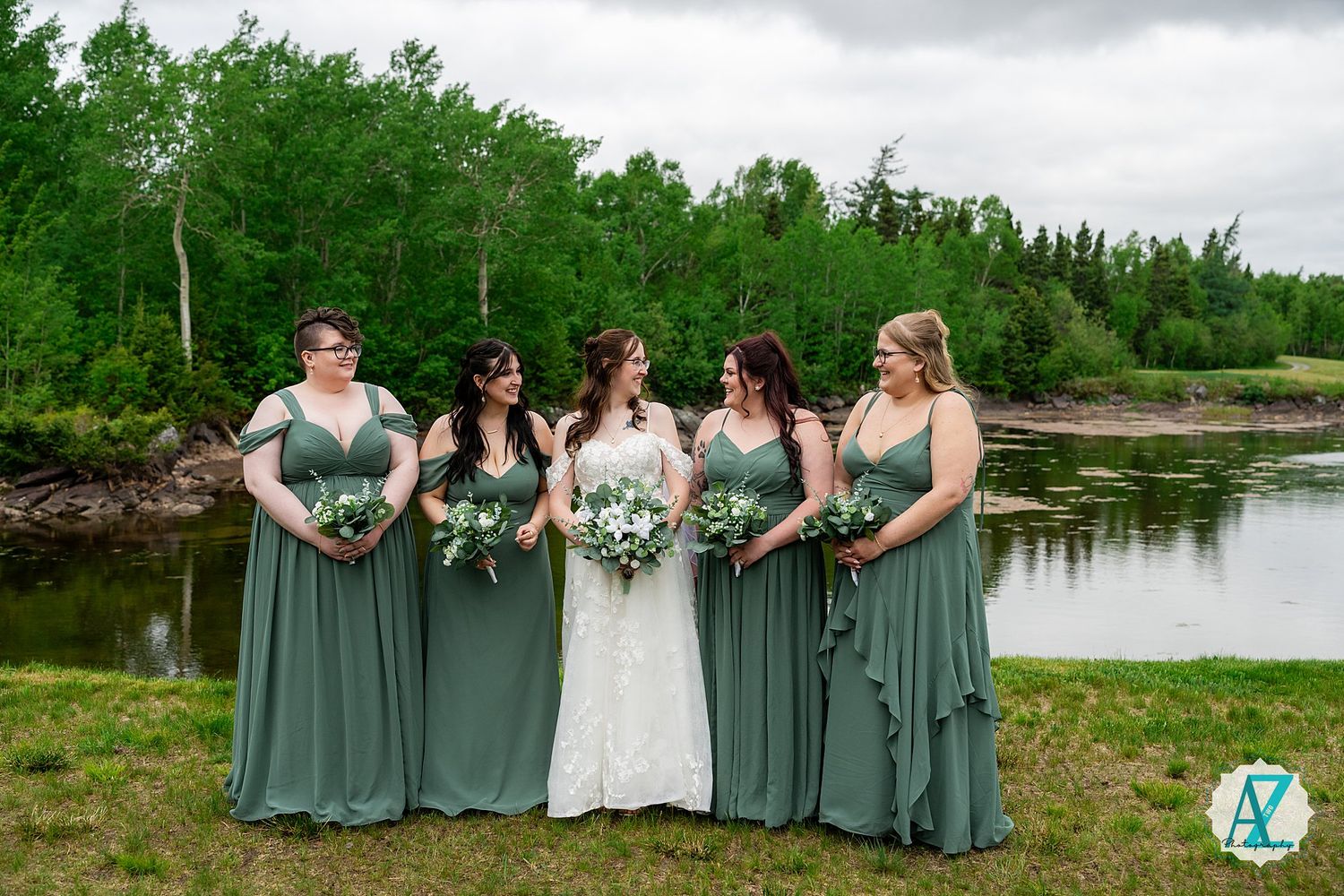 Bridal party in sage green dresses holding floral bouquets pose by serene waterfront on cloudy day.
