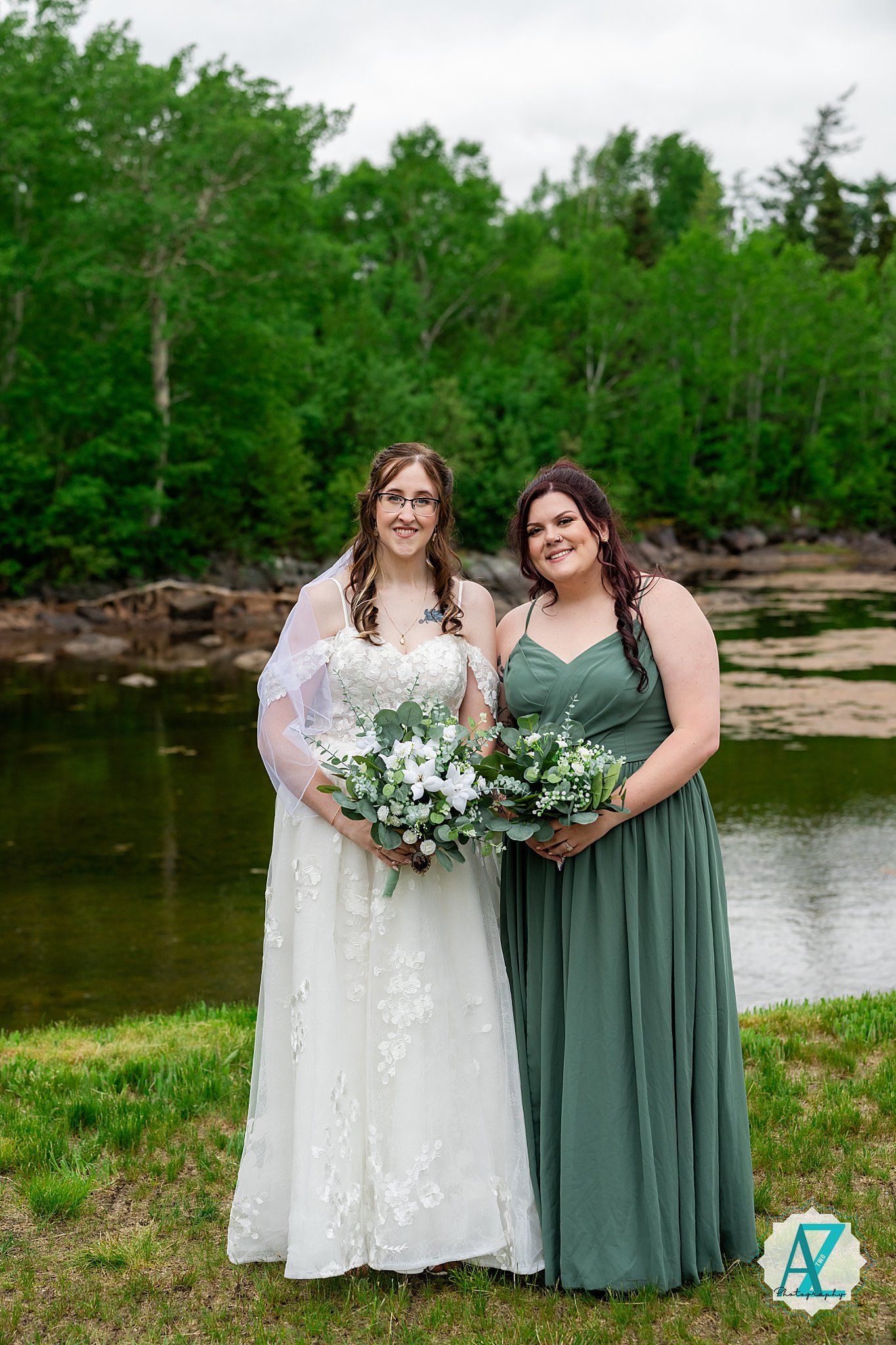 Two women in formal dresses stand by a lake with bouquets, wearing white and sage green gowns against green foliage.