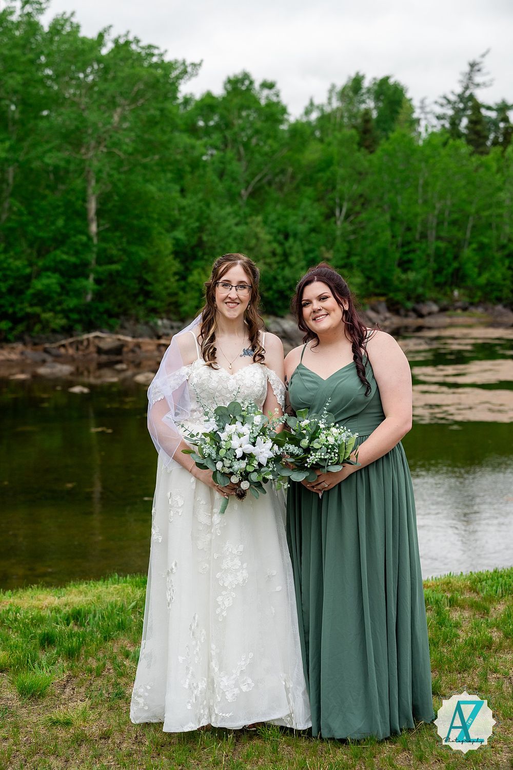 Two women in formal dresses stand by a lake with bouquets, wearing white and sage green gowns against green foliage.