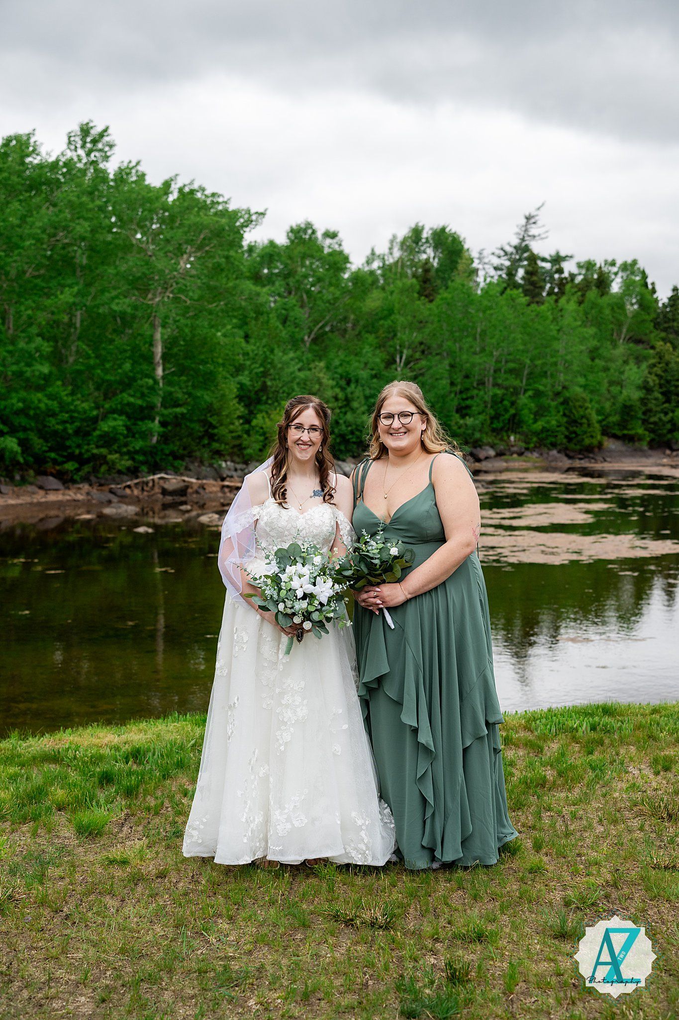 Two people in formal attire stand together by a serene lake with green trees in the background under cloudy skies.