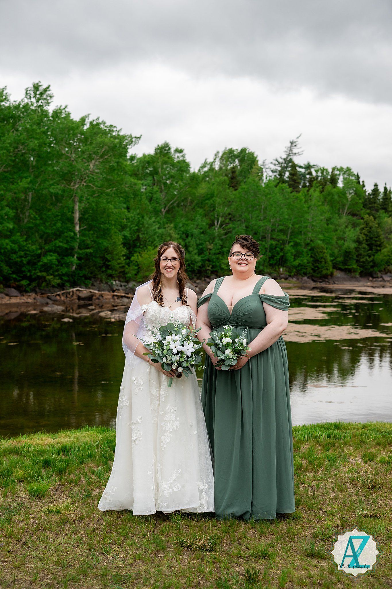Two people in formal gowns pose together at a waterfront wedding ceremony with lush green trees in the background.