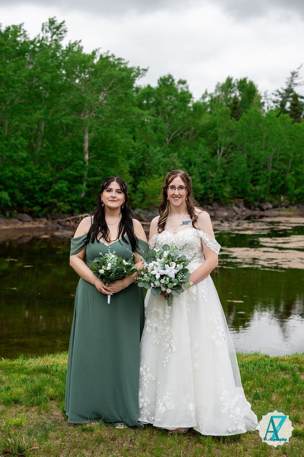 Wedding party stands together on grassy shore beside tranquil lake with lush forest backdrop.