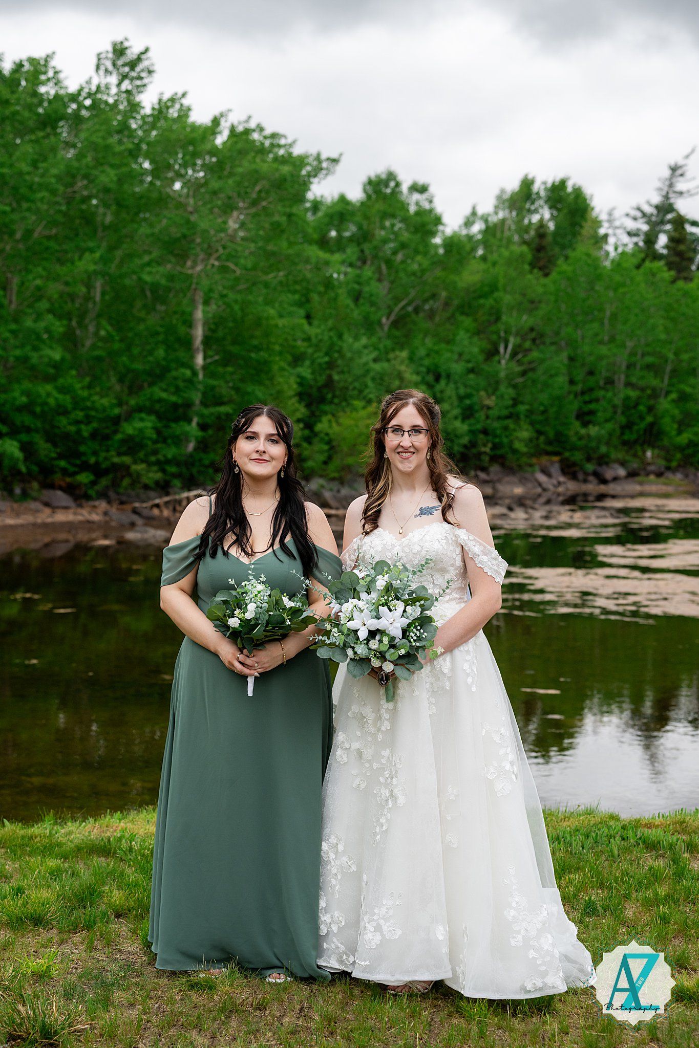 Wedding party stands together on grassy shore beside tranquil lake with lush forest backdrop.