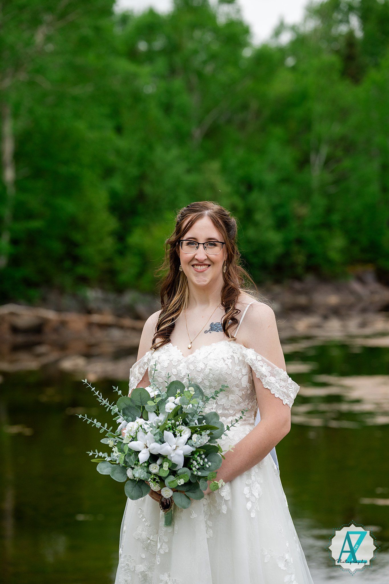 A bride in an off-shoulder lace wedding dress holds a white and green bouquet while standing by a serene pond.