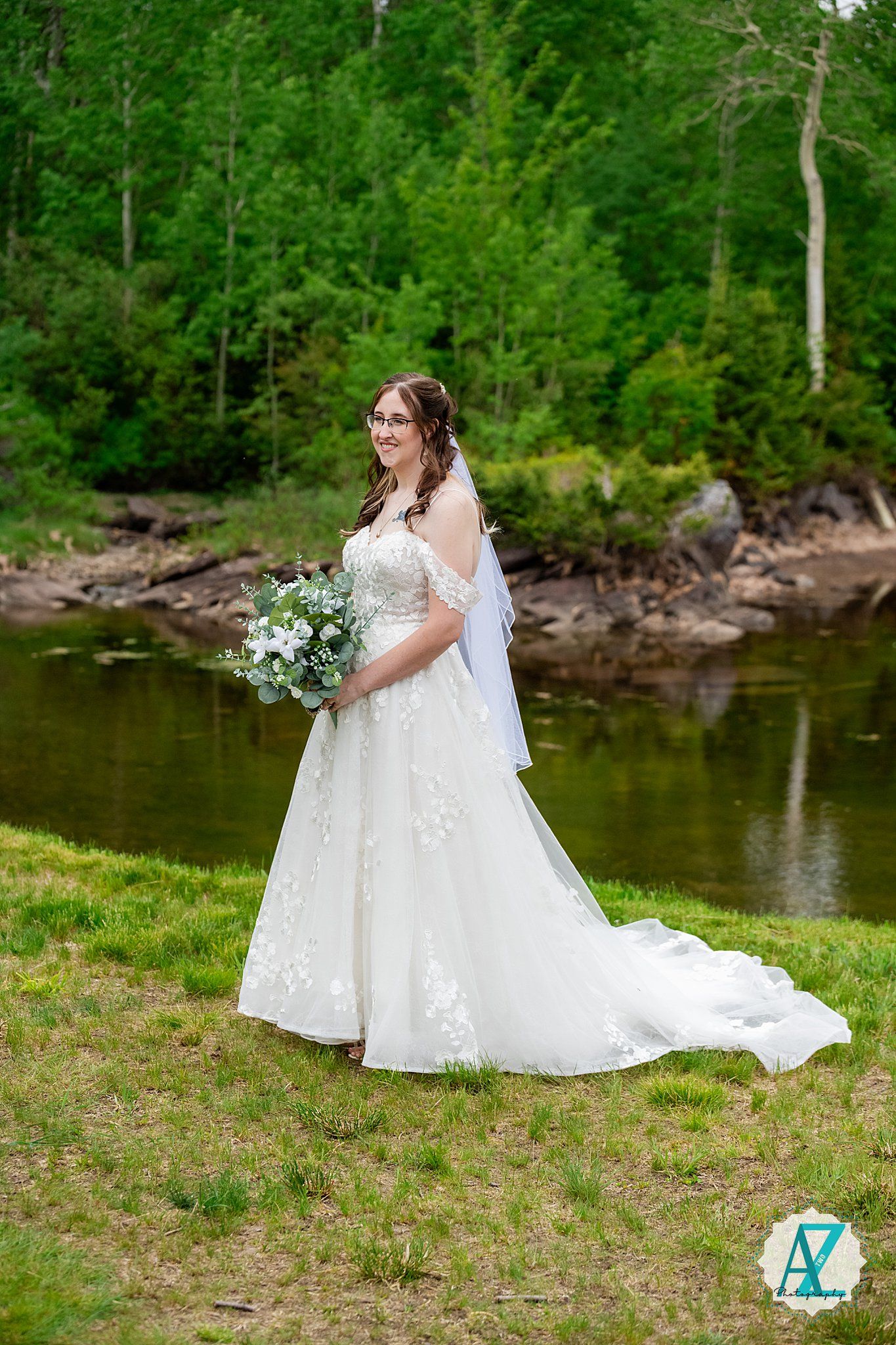 A bride in a white lace wedding dress sits gracefully by a serene lakeside surrounded by lush green forest.