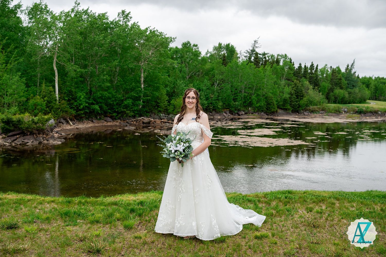 A white wedding dress stands out against a natural backdrop of green trees and tranquil water along a riverbank.