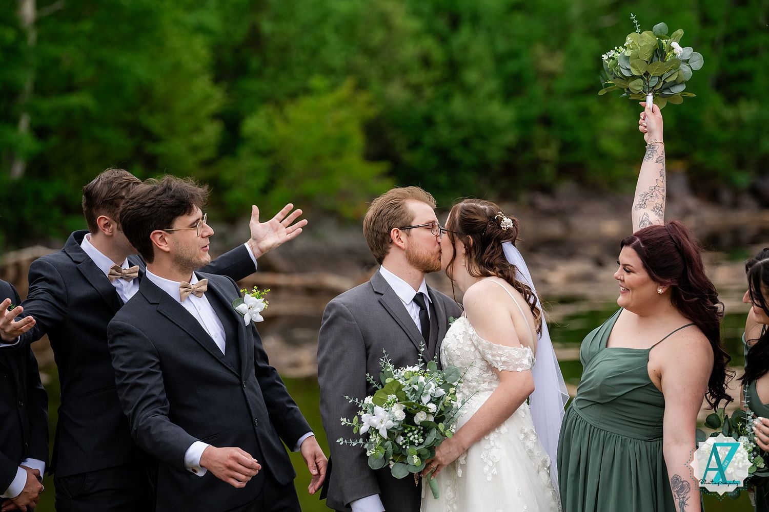 Wedding party looks on as couple shares a kiss during outdoor ceremony with green foliage and floral decor.