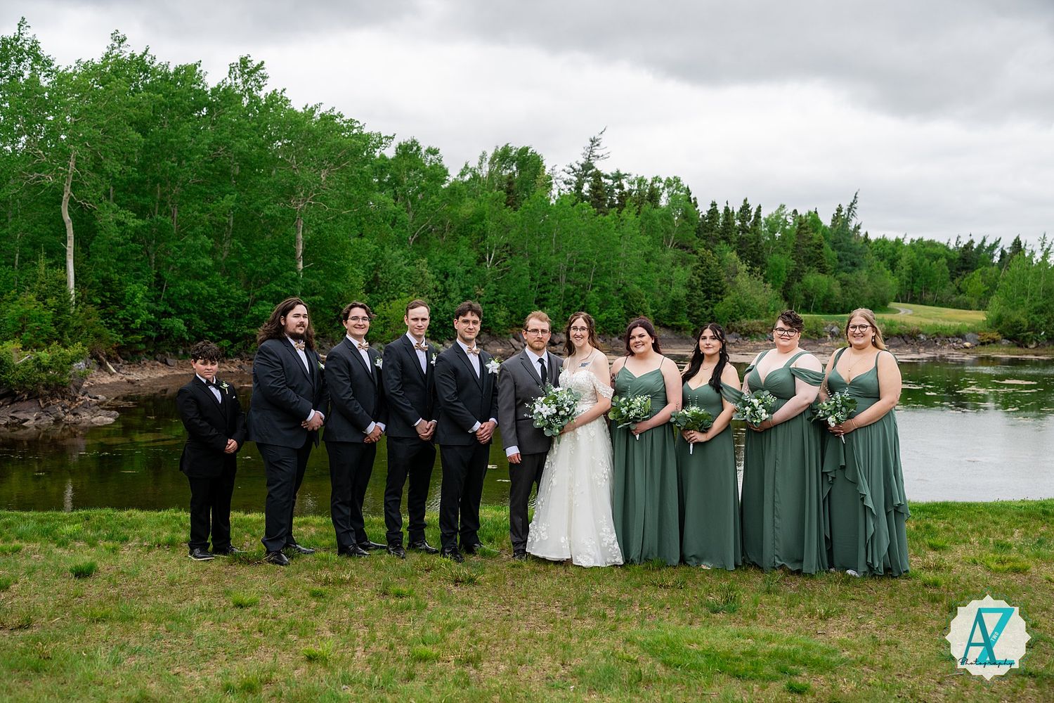 Wedding party stands in line by a scenic lake with bridesmaids in sage green dresses and groomsmen in dark suits.
