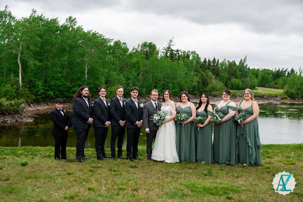 Wedding party stands in line by a scenic lake with bridesmaids in sage green dresses and groomsmen in dark suits.