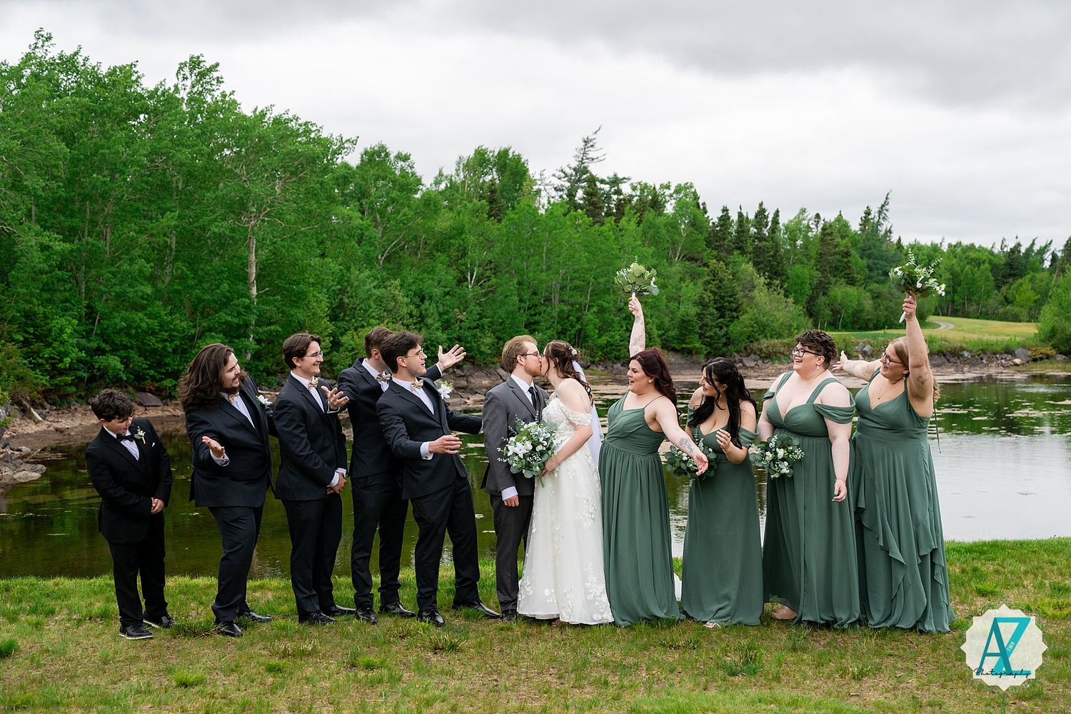 Wedding party stands outdoors by a lake with groomsmen in dark suits and bridesmaids in sage green dresses.