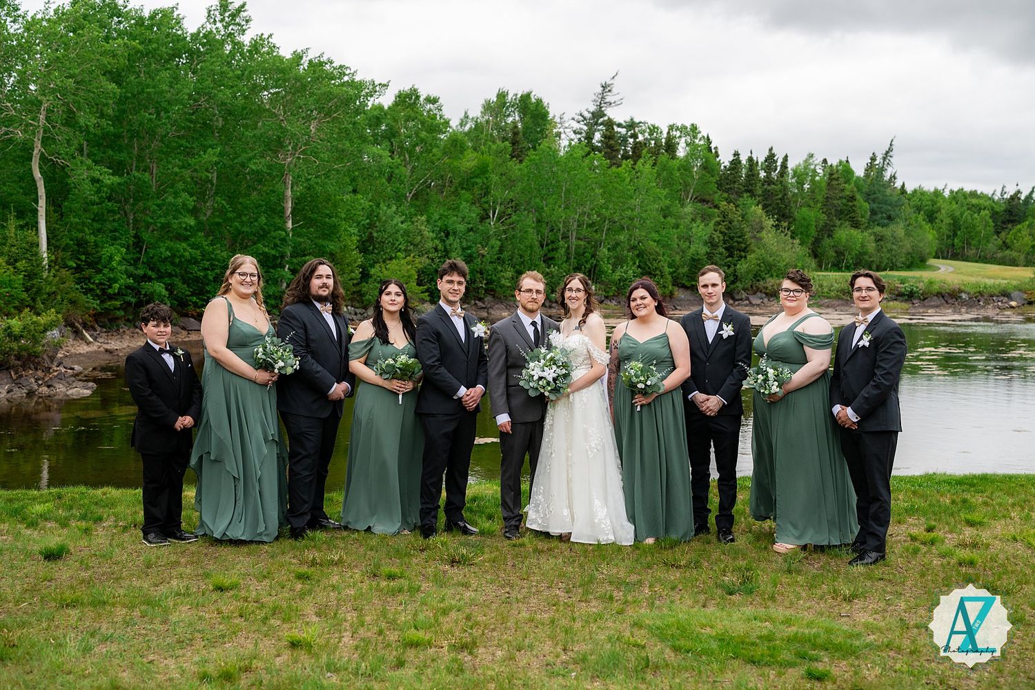 Wedding party group poses outdoors against scenic landscape with trees and water in the background.