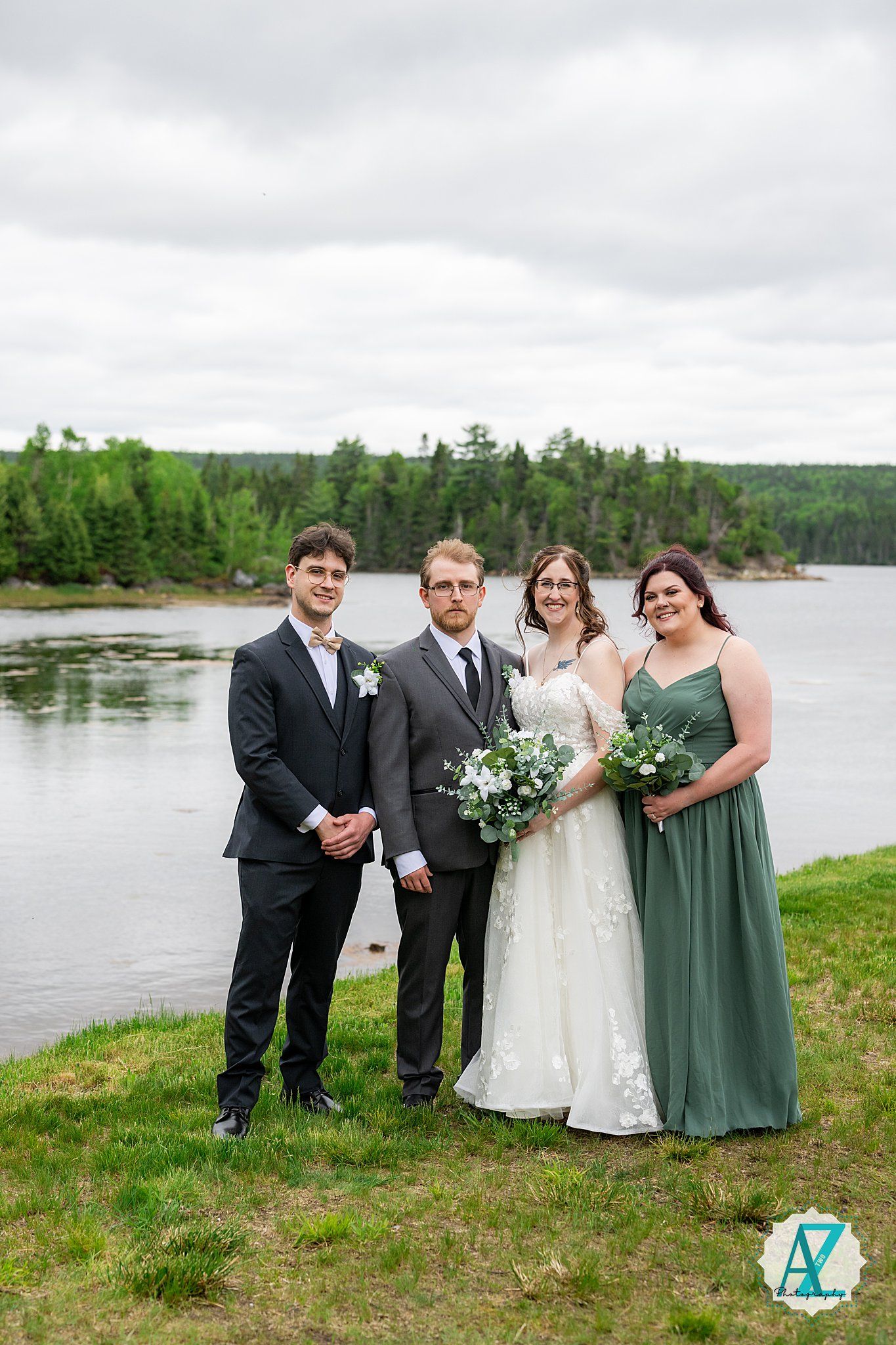 Wedding party in formal attire poses along scenic lakeside setting with green forest background.