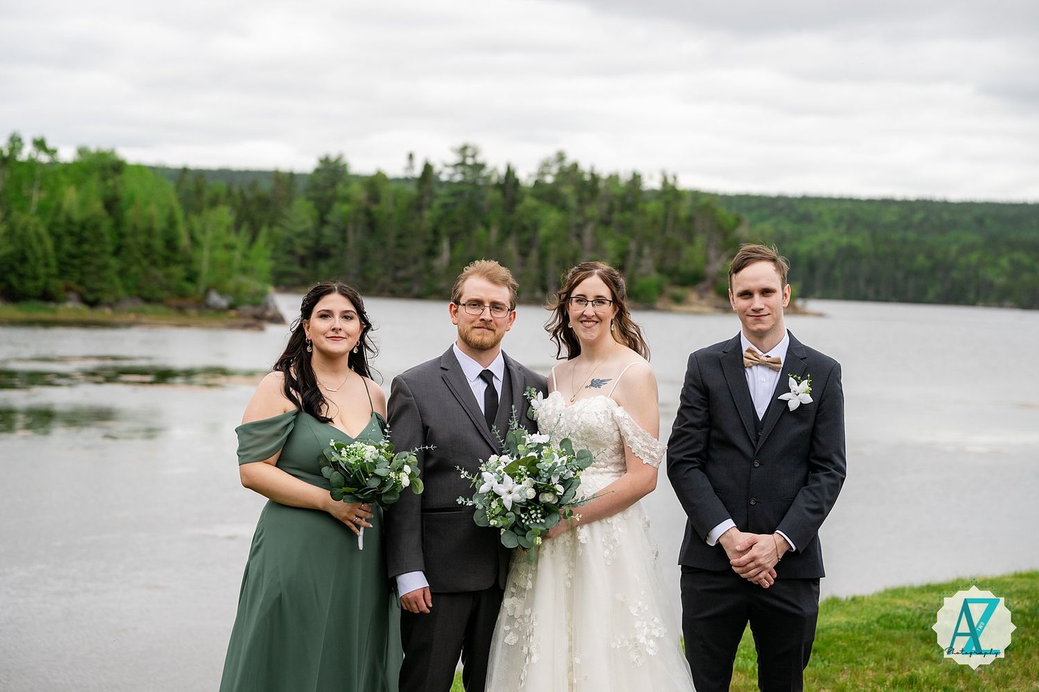Wedding party members in dark suits and sage green dresses stand together near a scenic waterfront.
