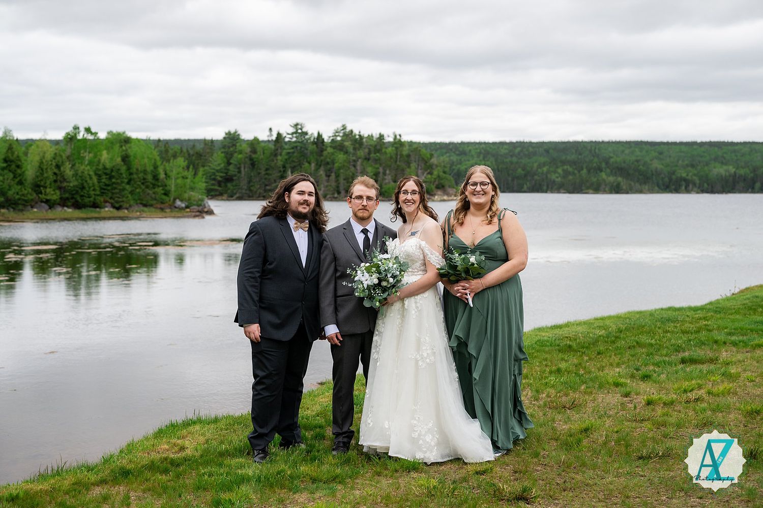 Wedding party stands on green lawn overlooking a serene lake with forested shoreline on an overcast day.
