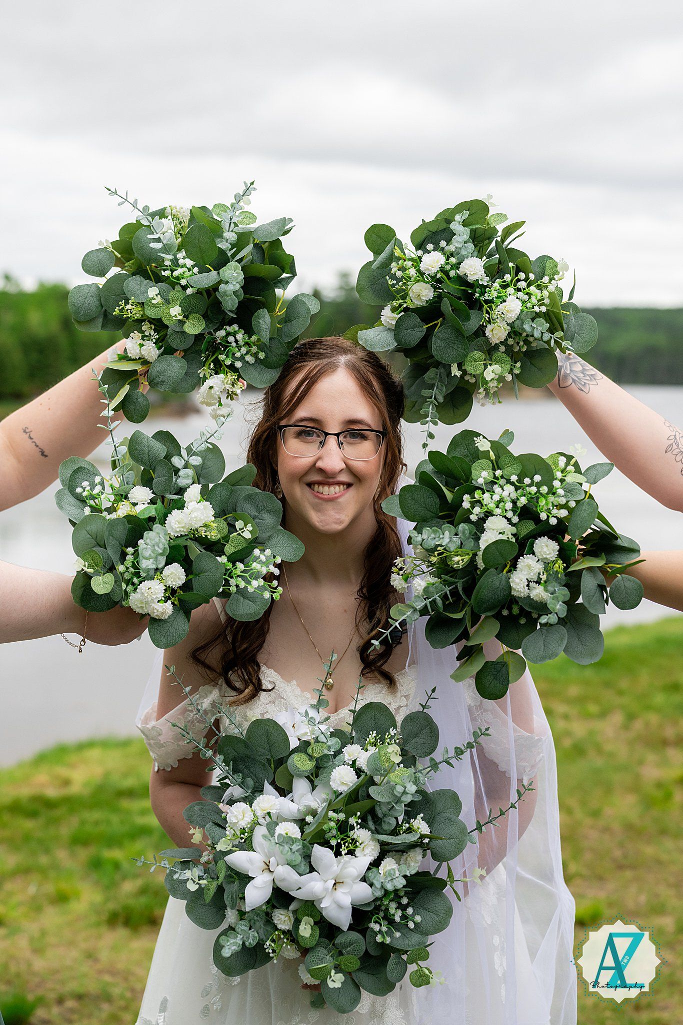 A bride in a white dress holds up five small floral arrangements of white flowers and eucalyptus against a cloudy sky.