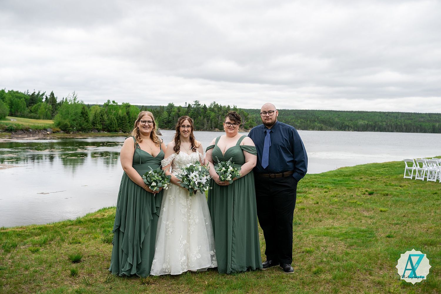 Wedding party stands together on grassy hill overlooking serene lake with forest backdrop under cloudy sky.