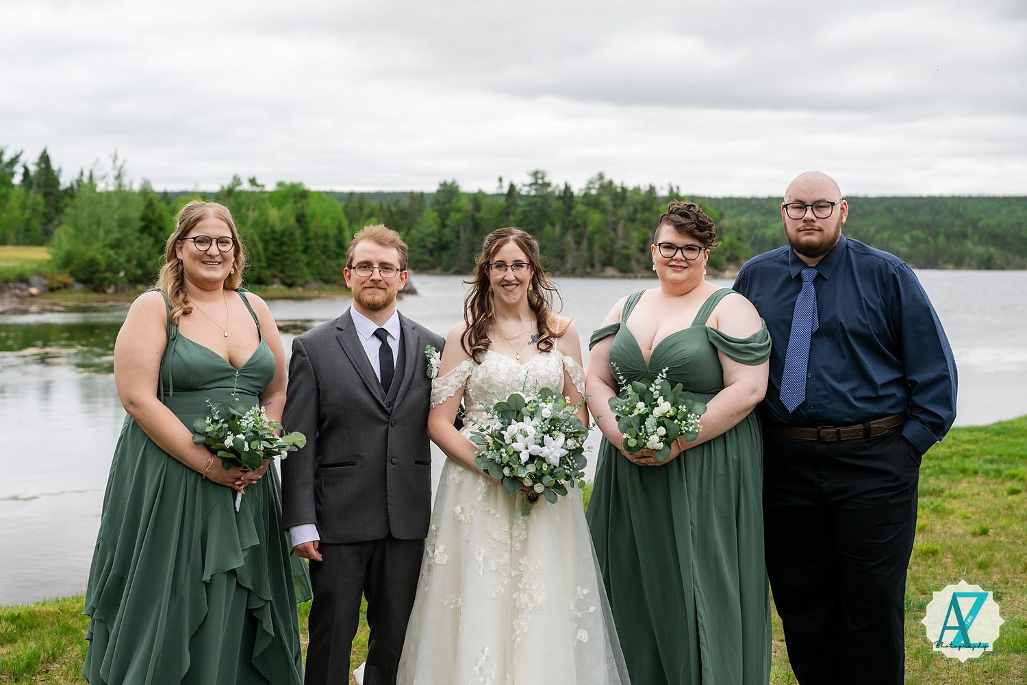 Wedding group poses on lakeside lawn with bridesmaids in sage green dresses and white floral bouquets.