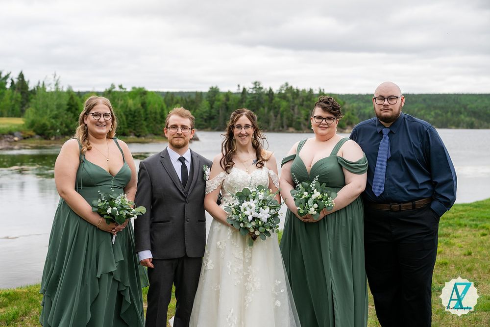 Wedding group poses on lakeside lawn with bridesmaids in sage green dresses and white floral bouquets.