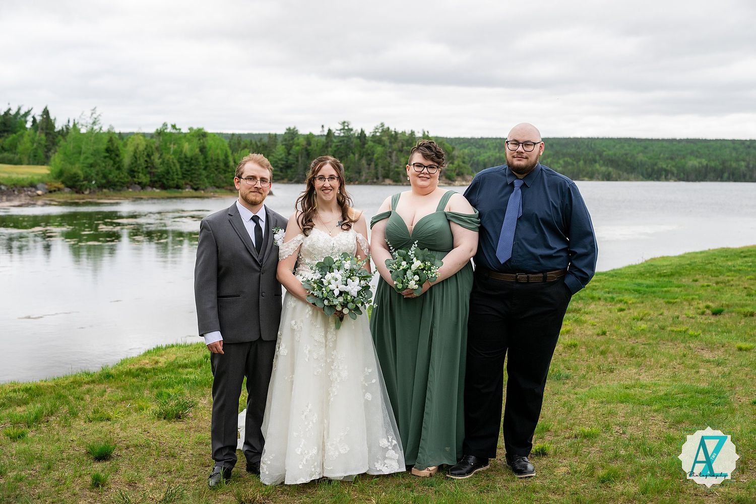 Wedding party of four stands together on a grassy hill overlooking a serene lake under cloudy skies.