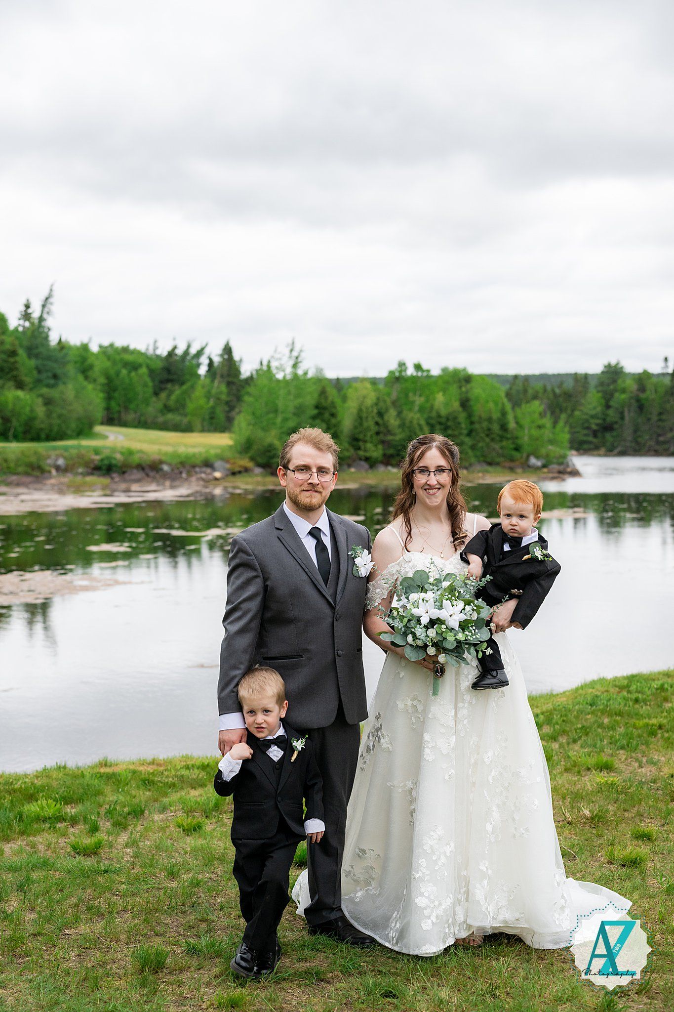 A wedding couple and two young children in formal attire pose together by a serene lake surrounded by green trees.