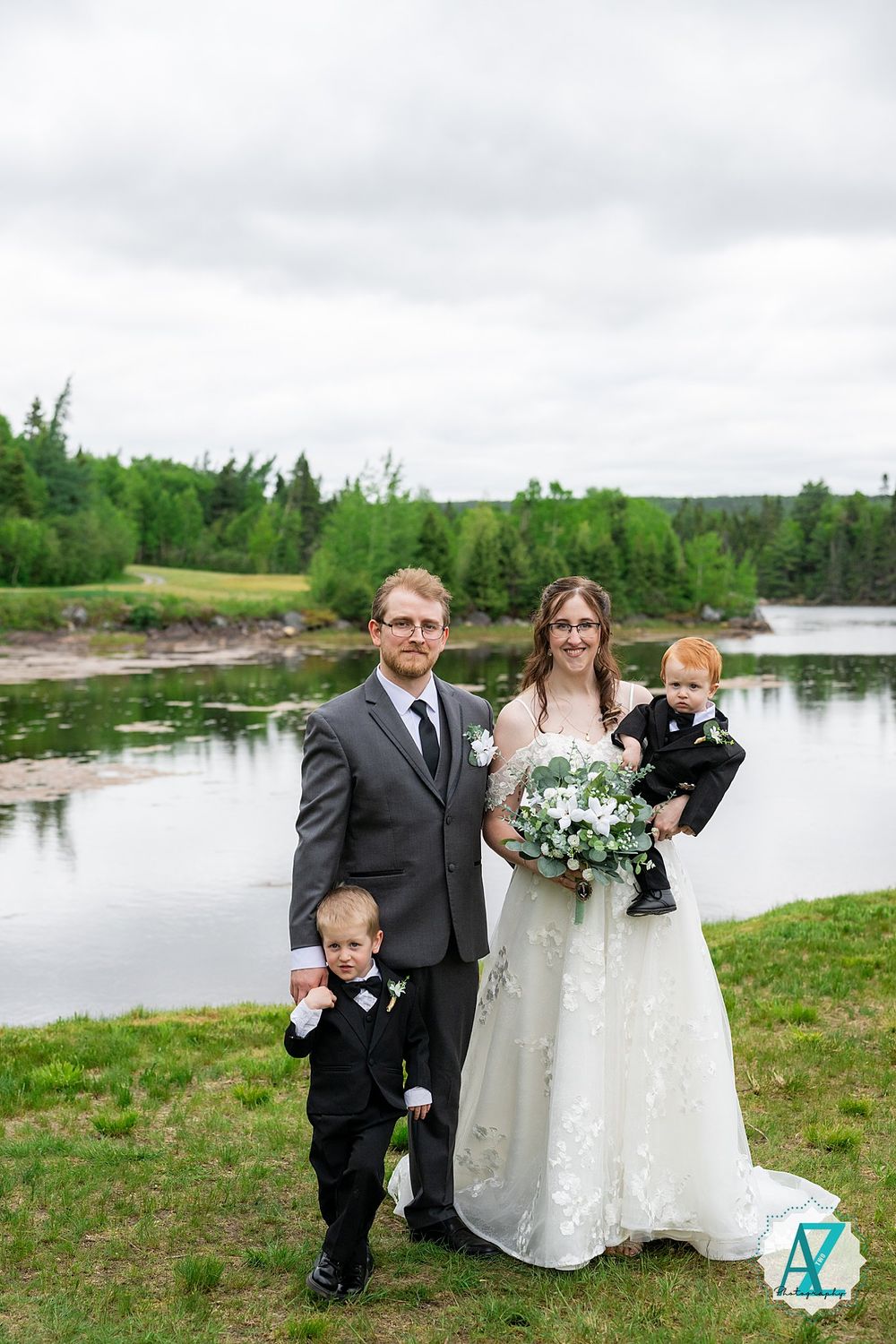 A wedding couple and two young children in formal attire pose together by a serene lake surrounded by green trees.