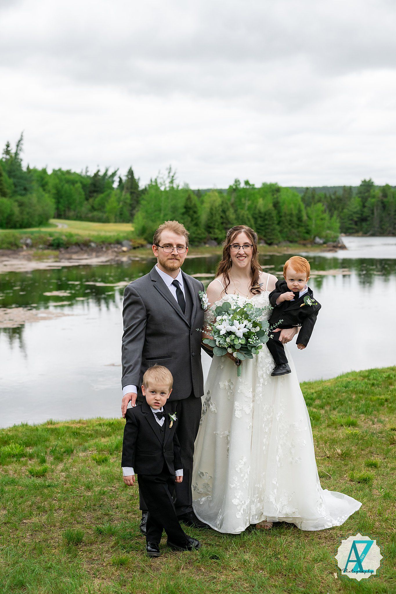 Wedding portrait of newlyweds with children at scenic waterfront location surrounded by trees and lush greenery.