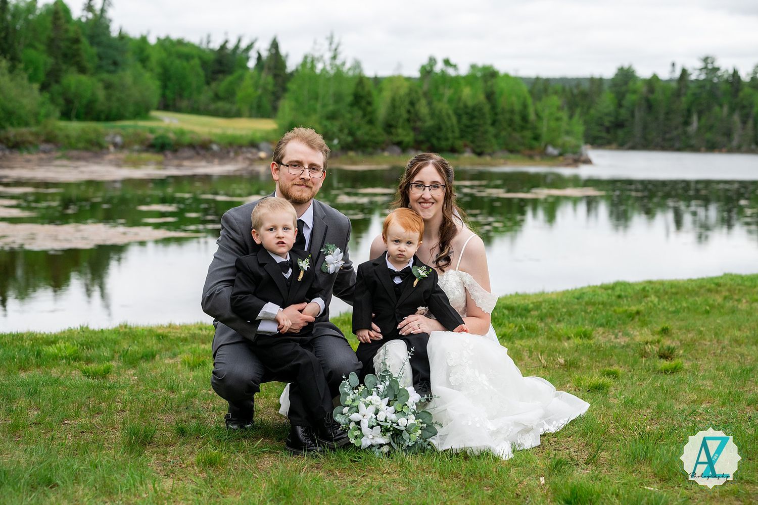 A newlywed couple with a dog poses by a serene lake surrounded by green forest on their wedding day.