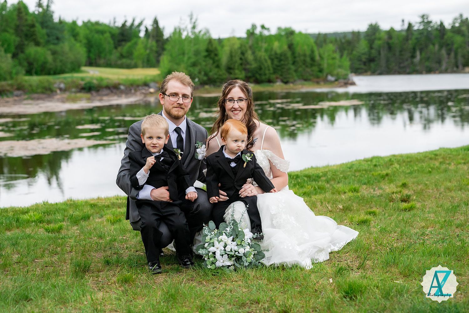 A family group poses together with their dogs by a tranquil lakeside on a wedding celebration day.