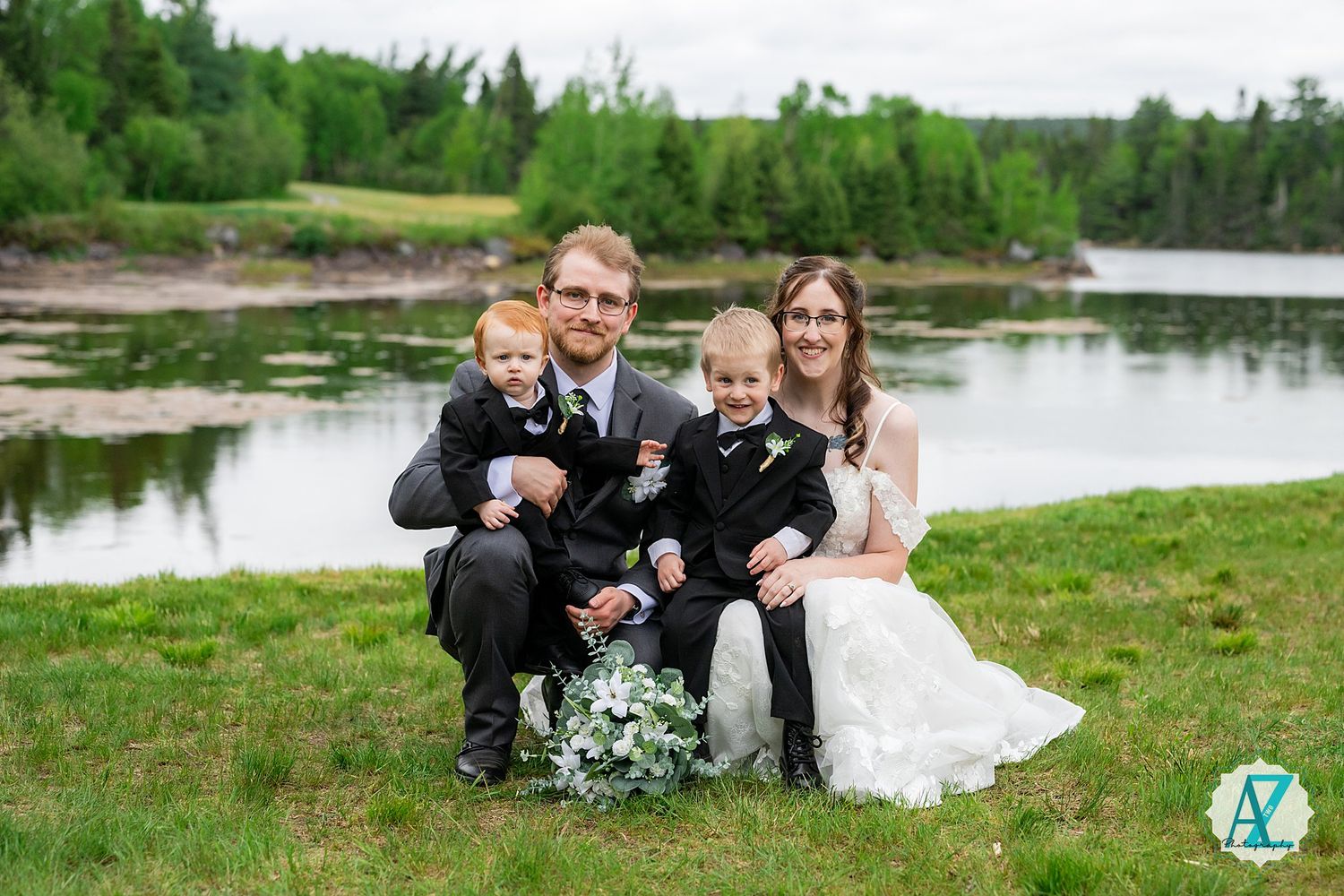 A wedding couple sits by a serene lake with greenery while holding bouquets of flowers on a grassy shore.