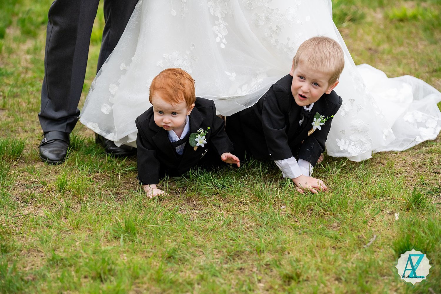 Two children in formal black suits playfully crawl on the grass next to a white wedding dress during an outdoor ceremony.