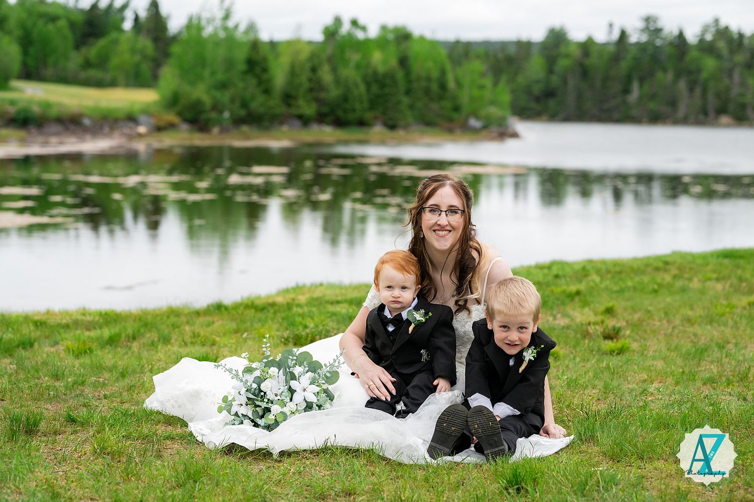 People sit together on a white blanket near a serene lake surrounded by green trees and grass on a bright day.