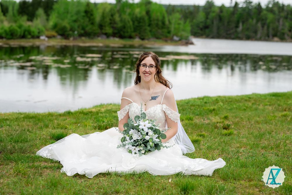 A bride in a white off-shoulder wedding dress sits on grass by a serene lake holding a blue and white bouquet.