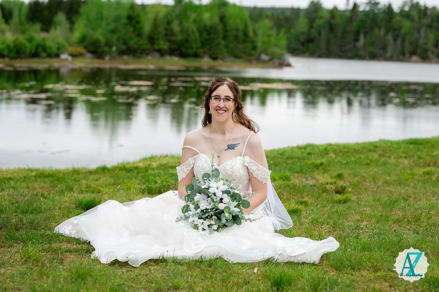 A bride in a white off-shoulder wedding dress sits on grass by a serene lake holding a blue and white bouquet.