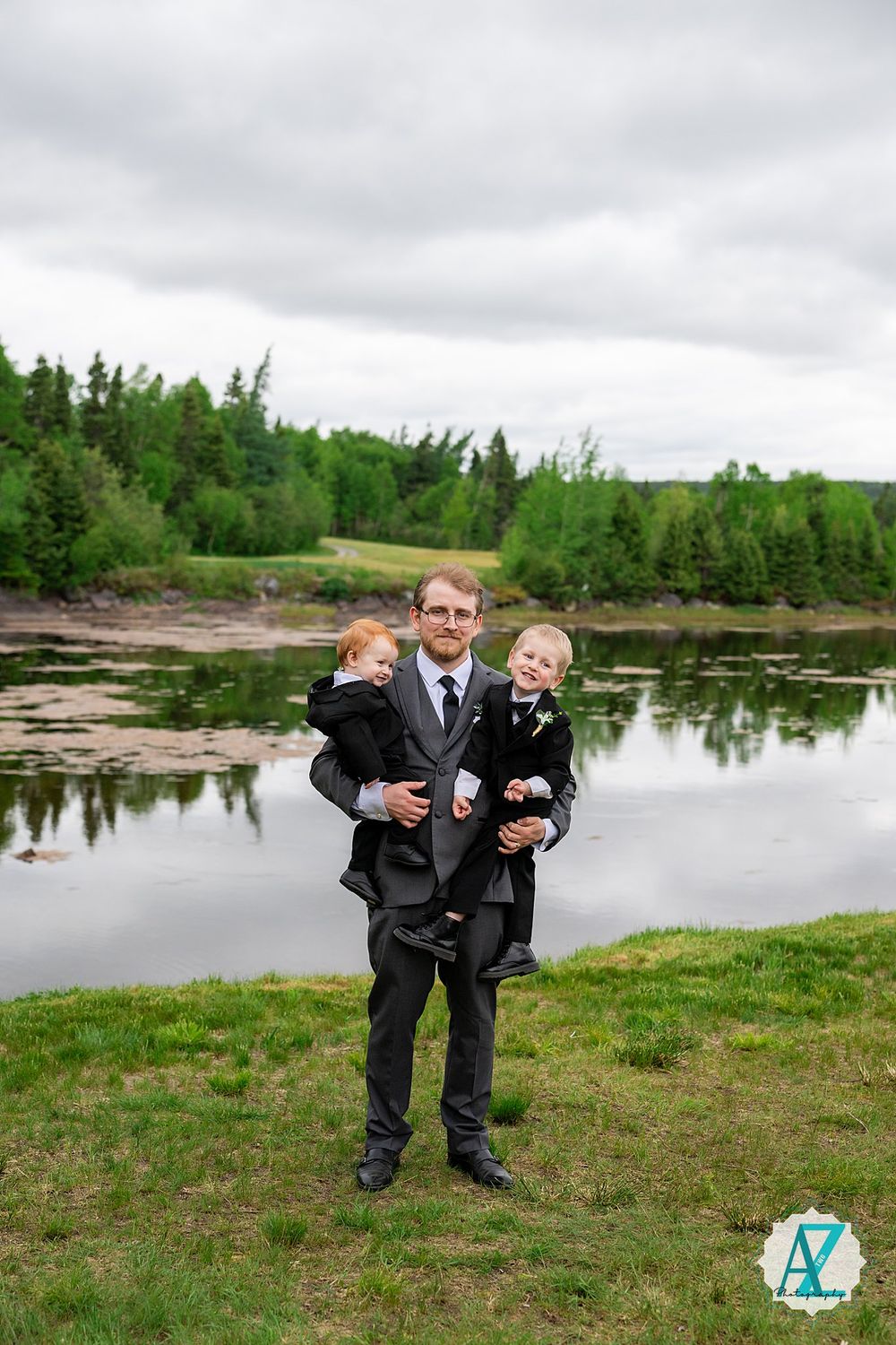 Family portrait taken by a tranquil lake with green trees and cloudy skies in the background.