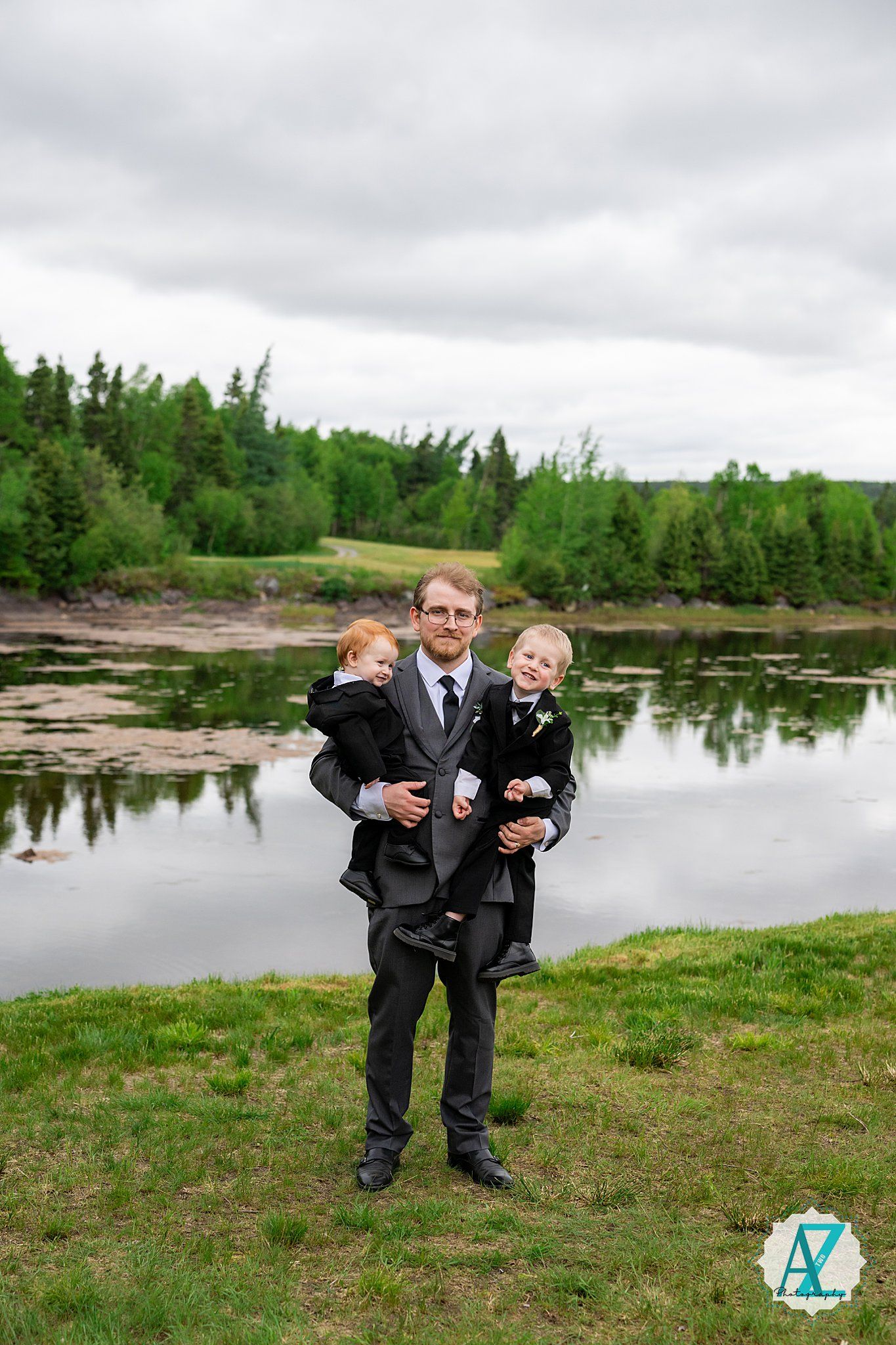 Family portrait taken by a tranquil lake with green trees and cloudy skies in the background.